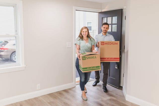 Couple moving into a new home, carrying boxes through the doorway, symbolizing the start of relocating to a new city.